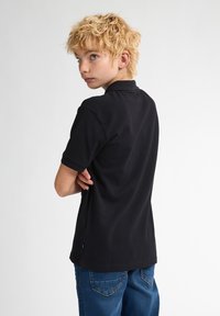 Svart pikétröja i bomullsblandning, med klassisk krage och korta ärmar. Matchad med blå denimjeans, visad bakifrån.