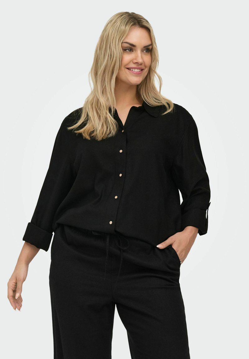 Lächelnde blonde Frau, die ein schwarzes Hemd mit hochgekrempelten Ärmeln und schwarze Hose trägt, eine Hand in der Tasche, vor einem schlichten Hintergrund.