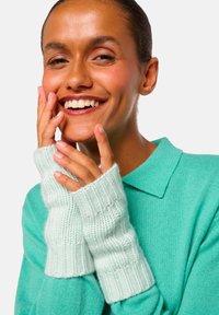 Femme souriante portant un pull teal et des gants sans doigts en tricot, de couleur vert menthe clair, touchant son visage avec les deux mains.