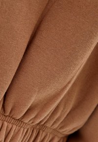 Närbild av brunt tyg med fin stickad textur och elastisk sammanknuten söm som skapar mjuka veck.