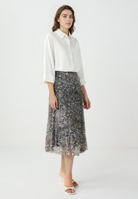 Weißes Button-up-Hemd mit langen Ärmeln, kombiniert mit einem plissierten Rock im Schlangenmuster in Erdtönen. Schlichte weiße Absatzsandalen.