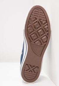 Semelle en caoutchouc d'une basket en toile bleue avec des accents blancs, présentant un motif de bande de roulement en forme de losange et un logo embossé. Design propre et minimaliste.