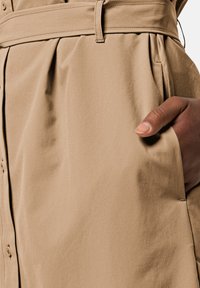 Beiges Kleid mit tailliertem Bund, gefälteltem Design und seitlichen Taschen. Der Stoff wirkt glatt mit einem leichten Glanz. Eine Hand ist in einer Tasche.