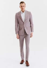 Jasnoróżowy garnitur o dopasowanym kroju, marynarka na dwa guziki, pasujące spodnie oraz biała koszula. Brązowe skórzane buty dopełniają całość.