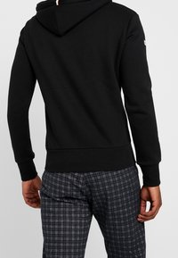 Uomo che indossa un hoodie nero e pantaloni a quadri grigio scuro, mostrato di spalle con le mani rilassate lungo i fianchi su uno sfondo semplice.