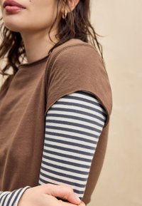 Bruine top met korte mouwen en een ronde hals, gelaagd over een grijs-wit gestreept shirt met lange mouwen. Het materiaal lijkt zacht met zichtbare stiksels.