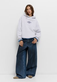 Felpa grigia con cappuccio e stampa "Sweet Dachshund Club", abbinata a jeans denim a gamba larga di un blu scuro. Le calzature presentano borchie decorative.