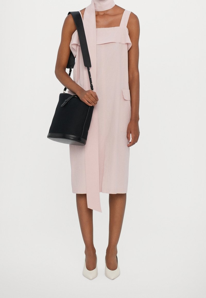 Vestido rosa sin mangas con detalle de volantes, dos bolsillos frontales y una larga bufanda. Bolso tipo cubo negro con una correa. Tacones blancos de punta.