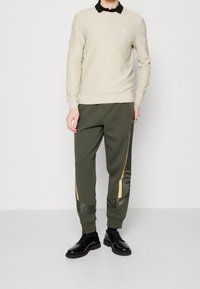 Suéter beige texturizado, camisa negra con cuello debajo; pantalones de chándal olivo con detalles brillantes en olivo y oro; zapatos negros con cordones.