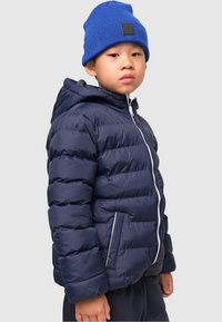 Marineblå pufferjakke med horisontalt quiltet design, hette og hvit glidelås. Kombinert med en blå strikket lue.