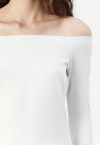 Top bianco a spalle scoperte con maniche lunghe realizzato in un tessuto morbido, con un design aderente e una consistenza liscia, caratterizzato da un ampio scollo.