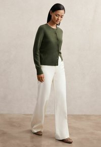 Zöld, kötött kardigán kerek nyakkivágással és gombos zárással, fehér, bő szabású nadrággal és barna balettcipővel párosítva.