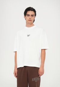 Bijela oversized majica od pamuka s kratkim rukavima, s malim crnim Reebok logotipom na prsima, u kombinaciji s smešnim trenerkama.