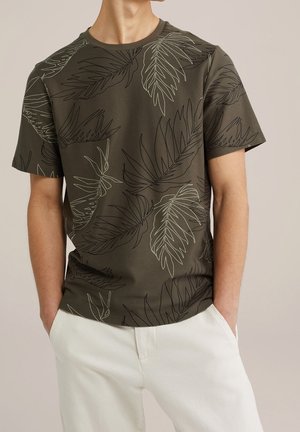 Mann trägt olivgrünes T-Shirt mit kurzen Ärmeln und schwarz-weißen tropischen Blattsilhouetten, kombiniert mit weißen Hosen, Hände in den Taschen.