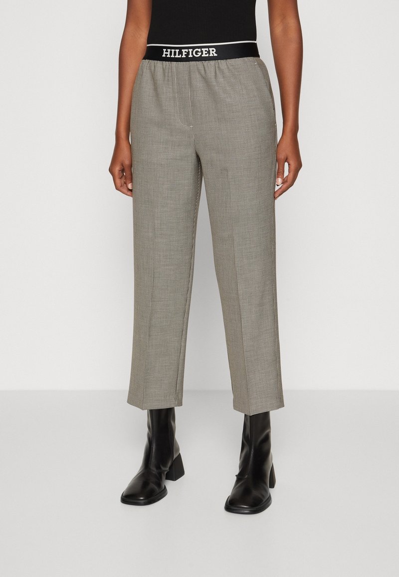 Harmaat ja mustat ruudulliset housut, joissa on vyötärönauhassa "HILFIGER"-logo, rento istuvuus ja lyhennetty pituus, yhdistettynä mustiin nilkkureihin.
