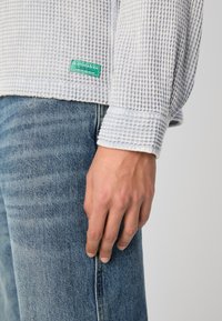 Vaffelstrukturert lys grå langermet skjorte med hvite mansjetter og grønn etikett. Kombinert med lyseblå denimjeans. Hånd som hviler på jeansene.