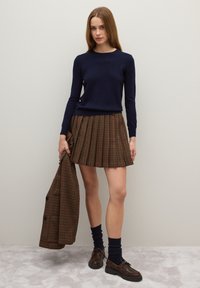 Marineblauer Pullover, plissierter Rock aus braunem Karo, knielang. Das Model hält außerdem einen passenden Karo-Mantel. Braune Schuhe mit dunklen Socken sind getragen.