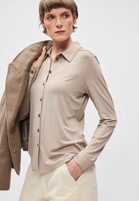 Bež košulja dugih rukava s vratom, koja ima dugmad sprijeda i glatku teksturu. Nosi se uz svijetlo obučenu kombinaciju.