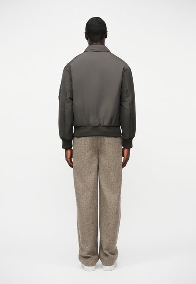 Homme de dos portant une veste bomber gris foncé, un pantalon large beige et des baskets blanches sur fond blanc.