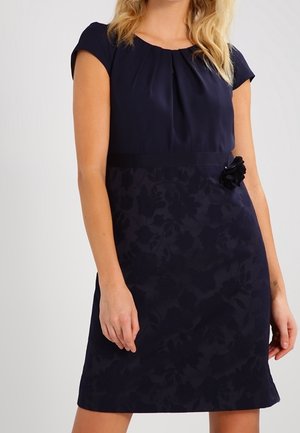 Kobieta ubrana w granatową sukienkę z krótkim rękawem, z kwiecistym wzorem na spódnicy i ozdobą w kształcie kwiatu w talii.