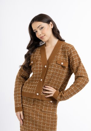 Junge Frau mit langen dunklen Haaren, die eine braune gemusterte Strickjacke und einen passenden Rock trägt, lächelt mit einer Hand in der Hüfte.