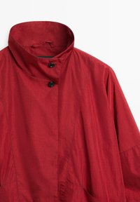 Veste légère rouge avec col montant et deux boutons noirs près de l'encolure, coutures visibles et coupe décontractée.