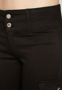 Μαύρο παντελόνι ψηλής μέσης με δύο μεταλλικά κουμπιά, εφαρμοστό, με λείο ύφασμα και λεπτομέρεια πλαϊνής τσέπης. Περιλαμβάνει ραφές κατά μήκος των ραφών.