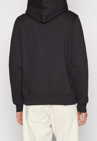 Black hooded sweatshirt with long sleeves, ribbed cuffs and hem, made of a soft material; worn over light-colored pants.