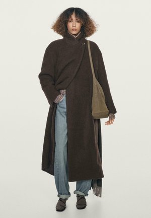 Donna che indossa un lungo cappotto marrone scuro, jeans azzurri arrotolati, scarpe marroni, mentre porta una borsa a tracolla verde oliva, in piedi contro uno sfondo semplice.