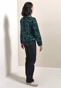 Giacca verde a motivo zigzag, jeans scuri e sneakers bianche; vista da dietro su uno sfondo neutro.