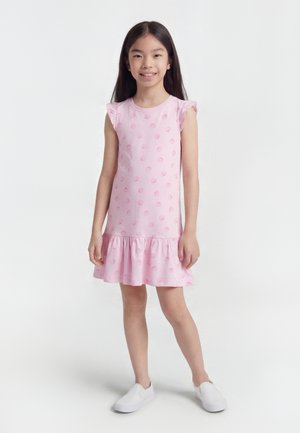 Niña joven de pie, vistiendo un vestido sin mangas rosa con estampados de conchas y zapatos blancos sin cordones, sonriendo frente a un fondo blanco liso.