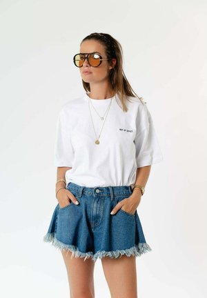 Femme portant un t-shirt blanc oversize avec le texte "Out of Office", un short en jean effiloché, de grandes lunettes de soleil teintées jaunes et des colliers superposés.
