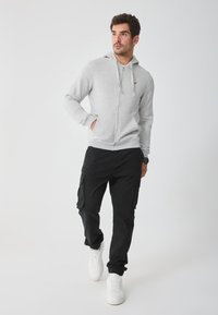 Sudadera con cremallera gris y capucha, con un pequeño logo. Combinada con pantalones cargo negros y zapatillas blancas con puños elásticos.