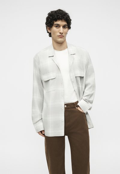 Νέος άνδρας με σγουρά μαλλιά που φοράει ένα ανοιχτό γκρι πουκάμισο με κουμπιά πάνω από ένα λευκό μπλουζάκι και καφέ παντελόνι, στέκεται μπροστά σε ένα απλό φόντο.