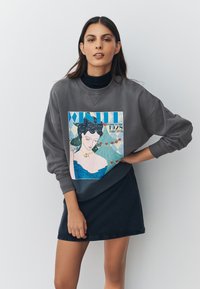 Harmaa ylipitkä collegepaita, jossa on värikäs grafiikka siniseen pukeutuneesta naisesta, jota ympäröivät sydämet ja geometriset kuviot. Yhdistetty tumman hameen kanssa.