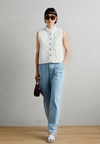 Colete acolchoado branco com colarinho mandarim, dois bolsos frontais e botões prateados, combinado com jeans azuis claros folgados e sandálias cinza claro.