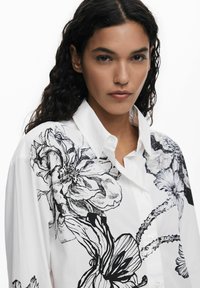 Camicia bianca con grandi illustrazioni floreali nere, design con bottoni, colletto a punta e maniche lunghe. Tessuto in cotone liscio.