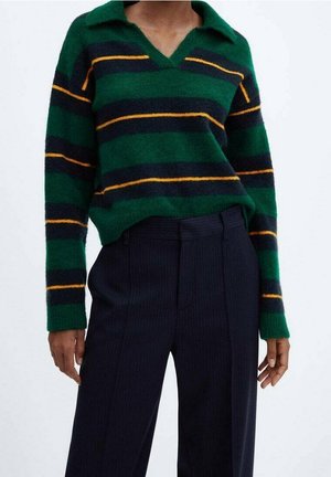 Personne portant un pull à manches longues rayé vert, marine et jaune, associé à un pantalon taille haute à rayures fines bleu marine foncé.