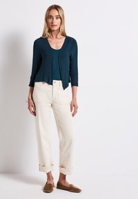 En teal lättviktskofta i lager över en matchande topp, parat med uppvikta krämfärgade byxor och bruna slip-on skor. Enkelt mönster, avslappnad stil.