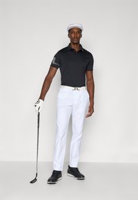 Camisola polo preta com mangas estampadas, calças brancas, cinto branco, sapatos pretos e um chapéu branco. Segurando um taco de golfe, de pé contra um fundo simples.