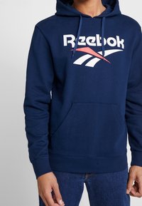Felpa blu navy con tasca a marsupio frontale, logo "Reebok" bianco e disegni accentati rossi e bianchi sul petto. Materiale in cotone.