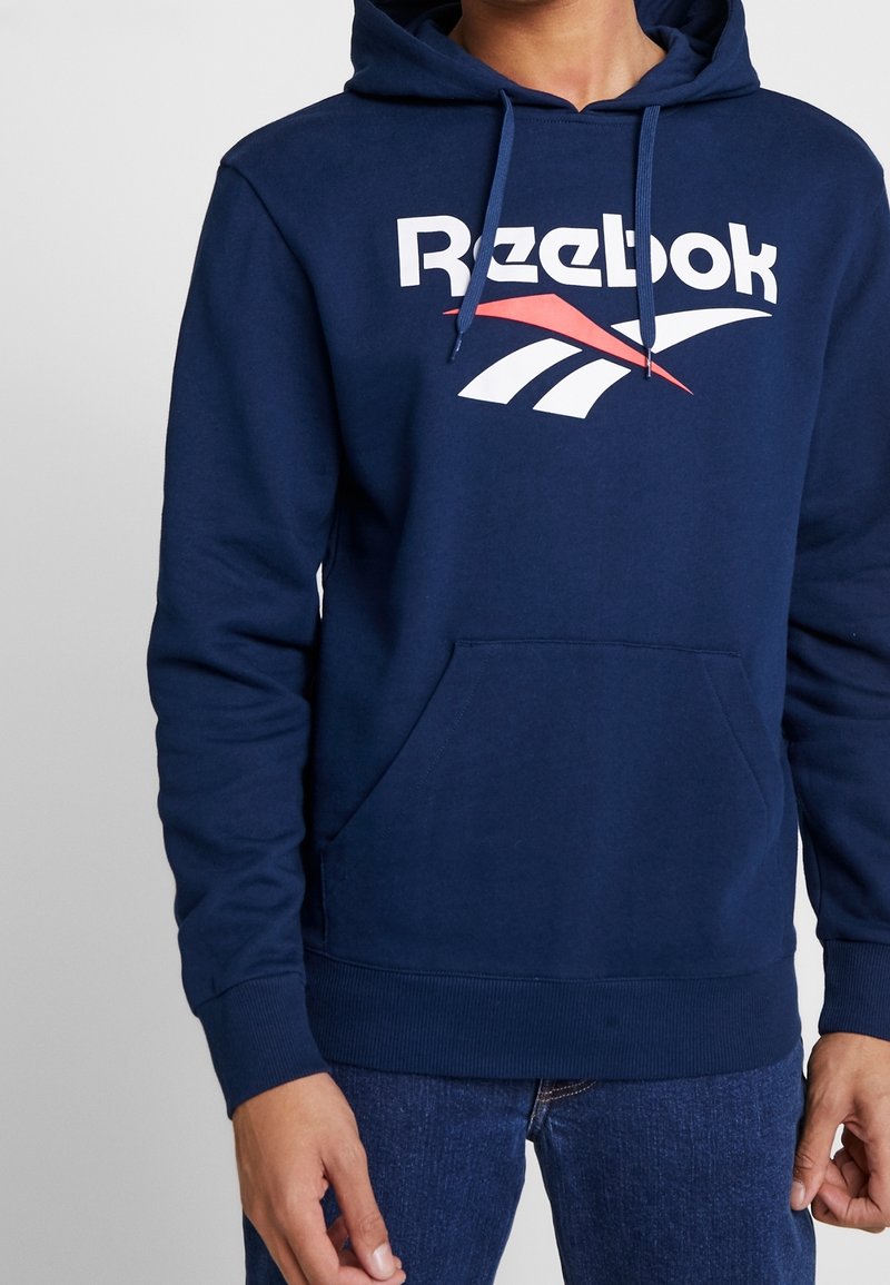 Felpa blu navy con tasca a marsupio frontale, logo "Reebok" bianco e disegni accentati rossi e bianchi sul petto. Materiale in cotone.