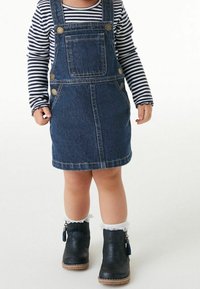 Kleinkind, das ein gestreiftes Langarmshirt, ein Denim-Overallkleid, weiße Rüschensocken und schwarze Stiefeletten trägt und auf einem weißen Boden steht.