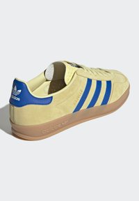 Tenisky Adidas Gazelle, vrchní část z jemné kůže v žluté barvě s modrými pruhy, měkký interiér, gumová podrážka, kulatá špička a texturované logo.
