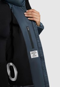 L'interno della giacca a vento blu presenta una fodera nera, una tasca con zip e un'etichetta che recita: "Un caldo abbraccio fatto di tessuto." Il numero "0" è visibile.