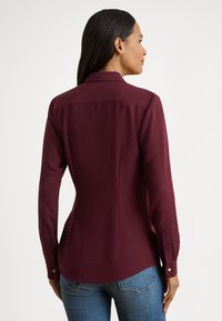 Bordo krāsas garām piedurknēm krekls ar apkakli, pieguļošu dizainu un pogām aizdarāmām aprocēm. Gluda auduma ar vieglu spīdumu, piegriezuma šuves uz muguras.