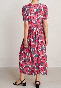 Kvinna stående barfota på ett betonggolv iförd en vit midiklänning med röd och grå blommönster och korta puffärmar.