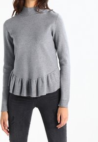 Maglione grigio a maglia con un collo alto e un orlo a balze a peplo. Maniche lunghe e texture liscia, abbinato a jeans scuri.