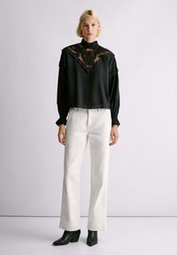 Blusa nera con yoke in pizzo, maniche a volant, abbinata a jeans bianchi a gamba dritta e stivaletti verdi con tacco e finitura texturizzata.