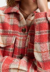 Rote und beige karierte Jacke aus strukturiertem Wollstoff. Besitzt große Knöpfe und Brusttaschen, hat einen Kragen und weichen Stoff.